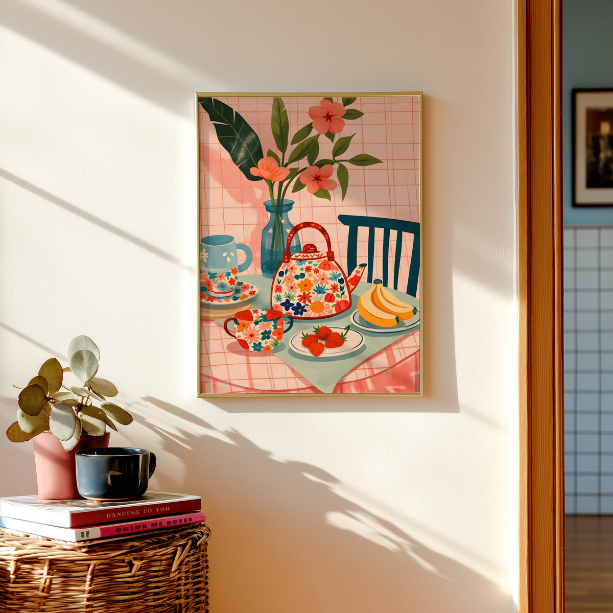 Floral teapot print in a gold frame above a basket of books and a pilea plant