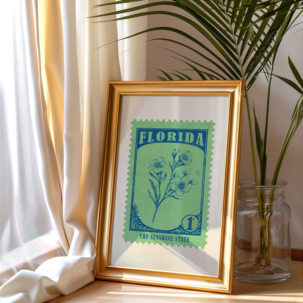 Florida stamp print in a gold frame leaning by a cream curtain with a palm in a glass jar