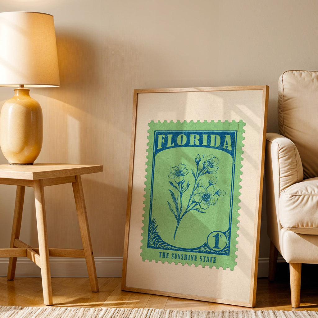 Florida stamp print in a wood frame resting on the floor beside a beige armchair and a yellow lamp