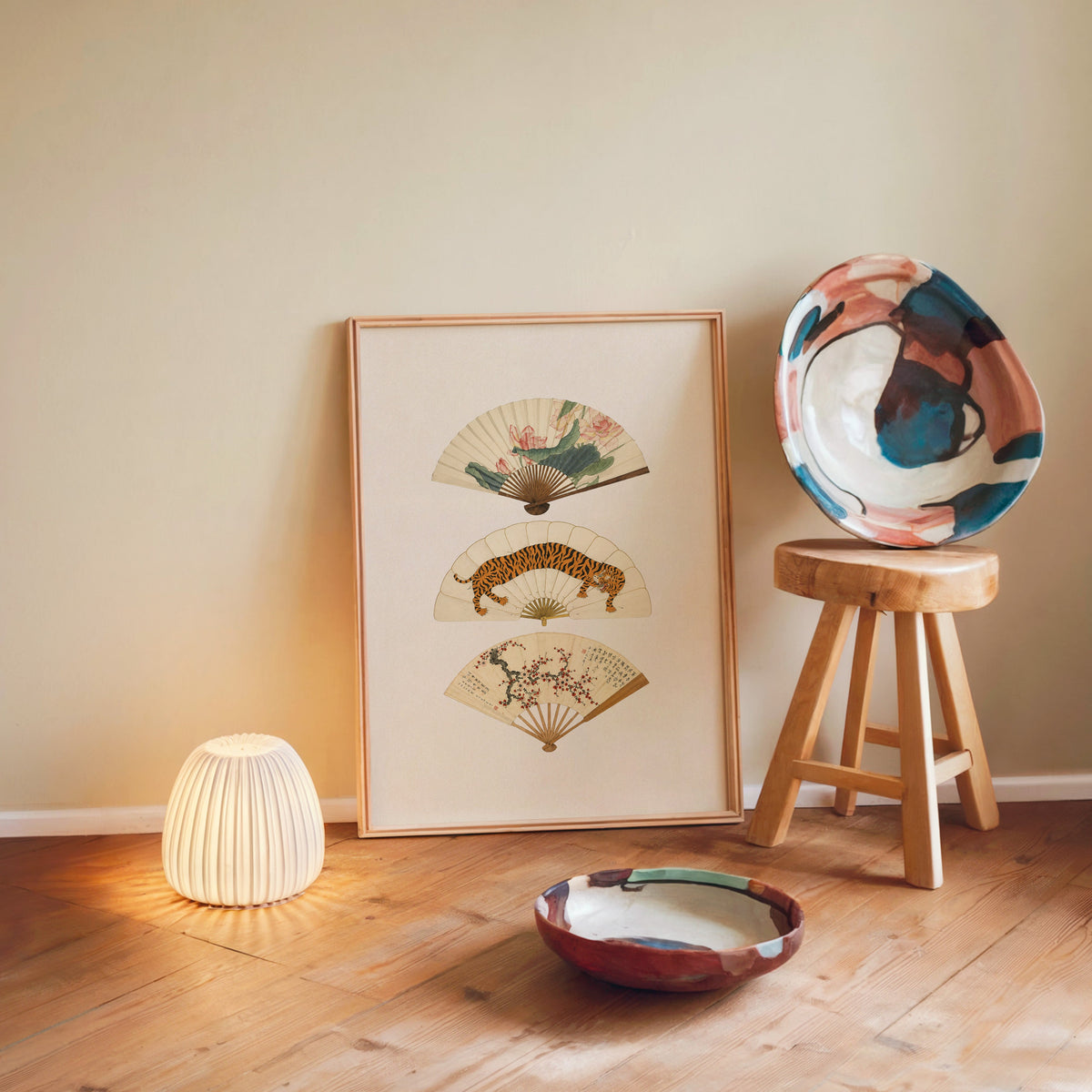 three painted hand fans print in a light wood frame leaning on a wooden floor by a glowing lamp and stool