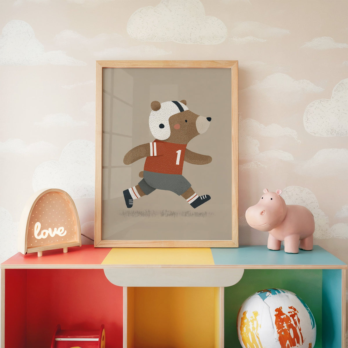 Football bear number one nursery print in an oak frame on a rainbow toy shelf against cloud wallpaper