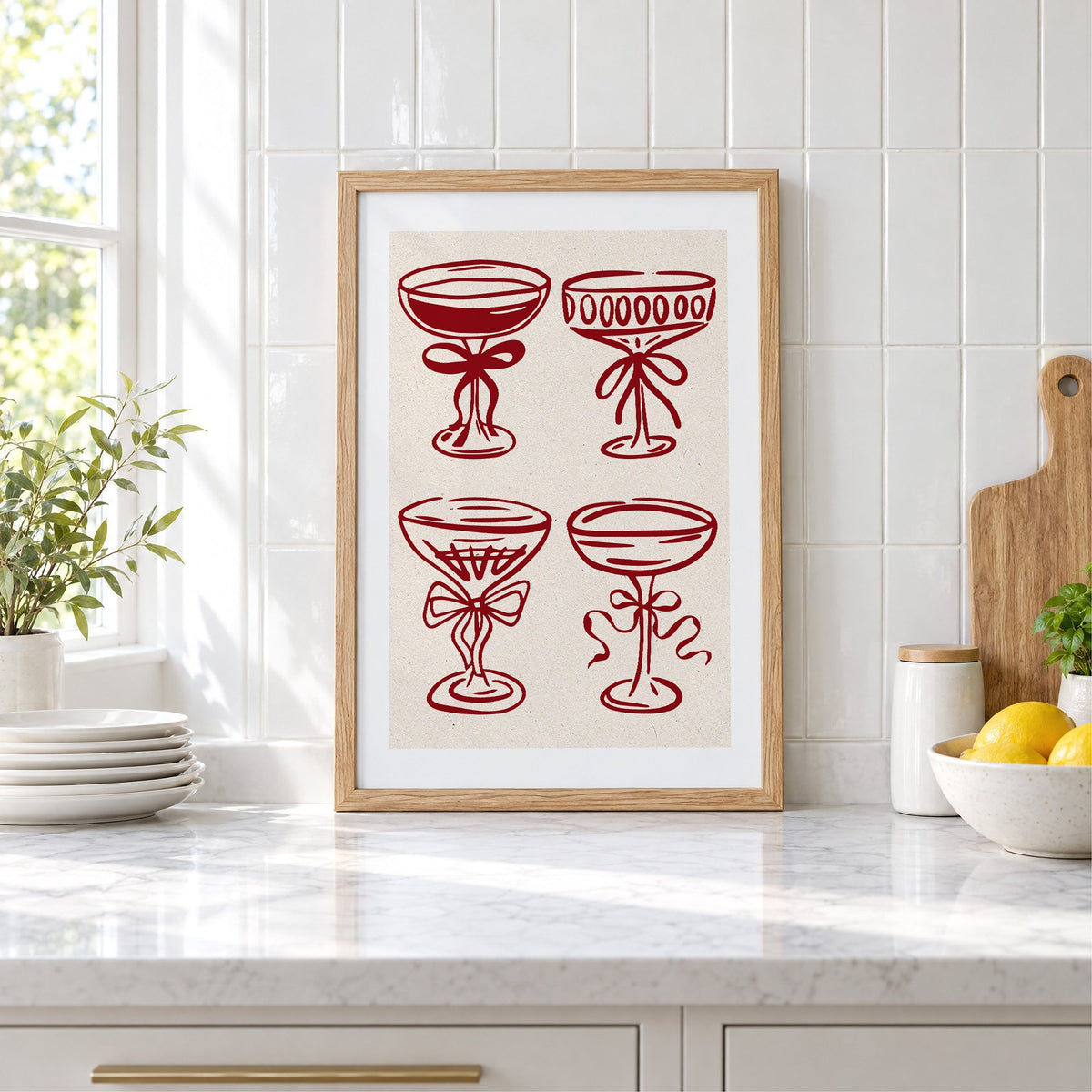 Four red coupe glasses with bows print in an oak frame leaning on a marble counter by white tiles and lemons
