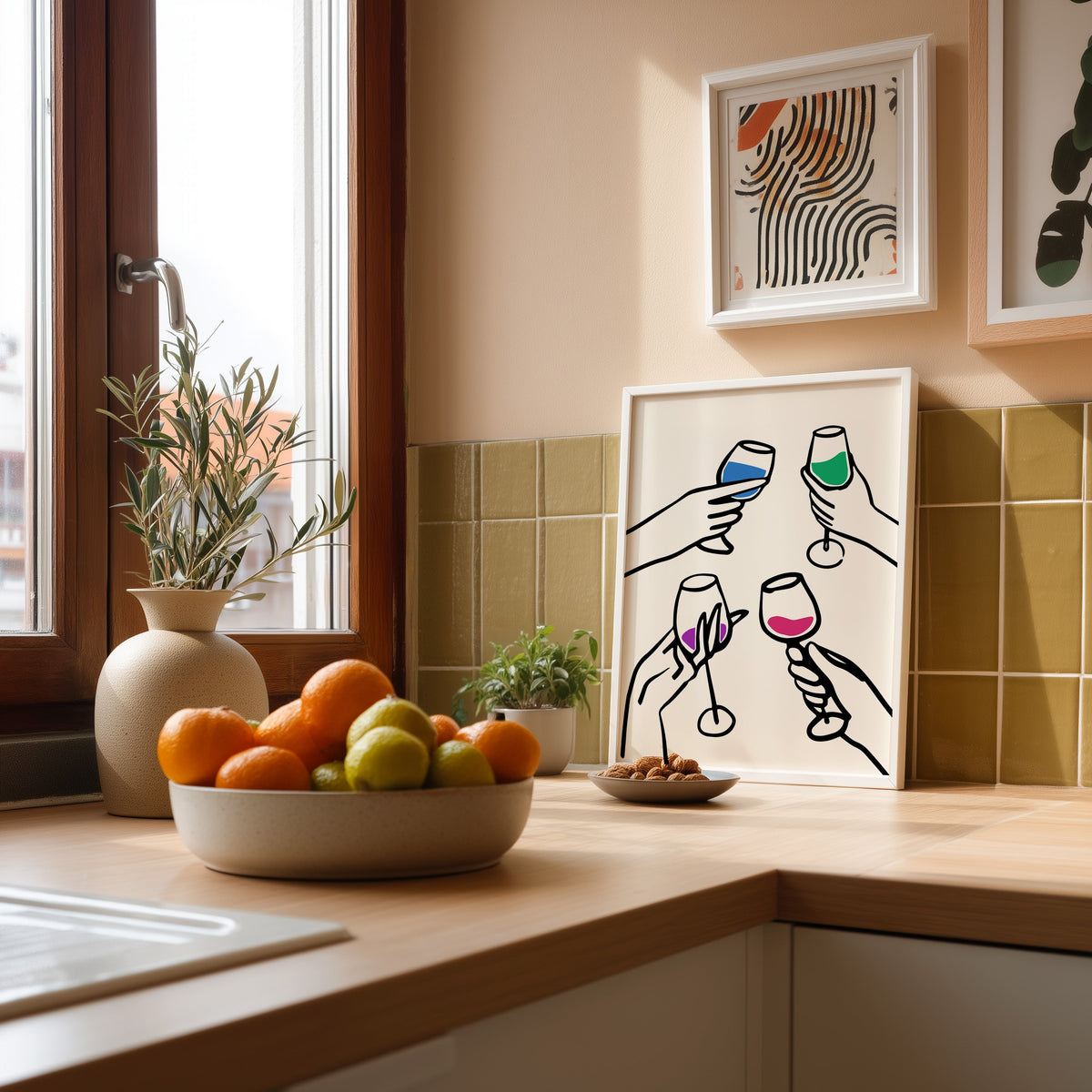 Four hands cheers colour pour print in a white frame leaning against olive green kitchen tiles beside a bowl of citrus
