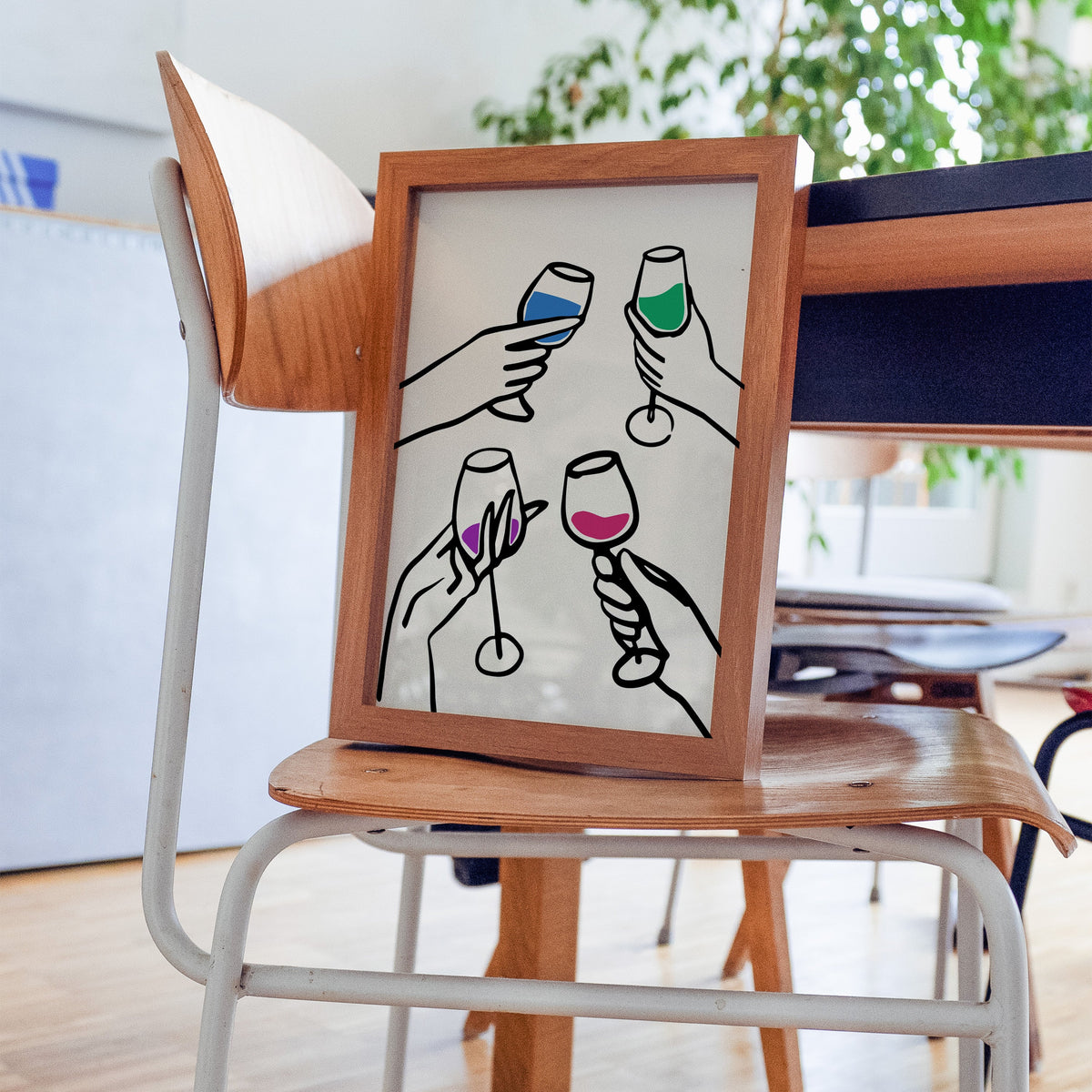Four hands cheers colour pour print in a walnut frame resting on a plywood studio chair beside a dark desk