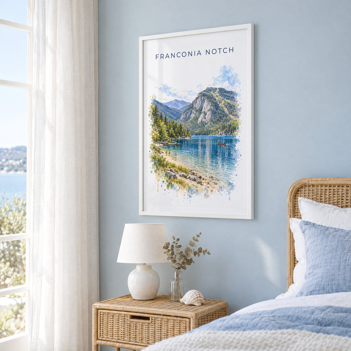 Franconia Notch watercolor print in a white frame on a pale blue wall beside a rattan bed in a coastal bedroom