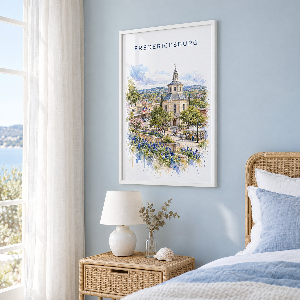 Fredericksburg, Texas watercolor print in a white frame on a pale blue wall beside a rattan bed in a coastal bedroom