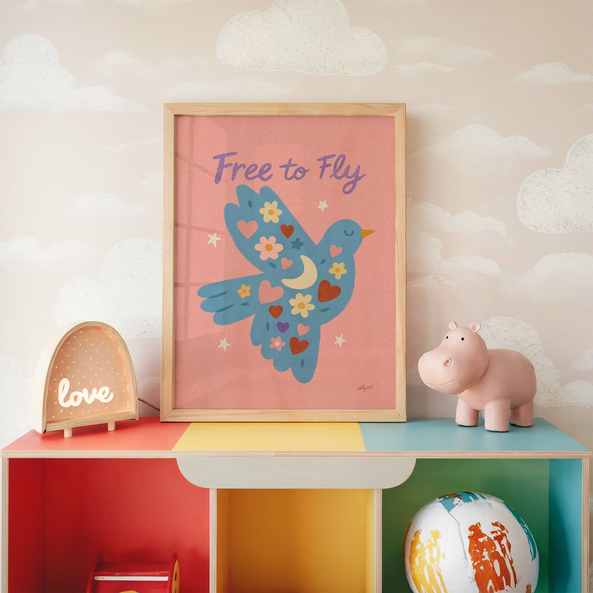 Folk blue bird free to fly nursery print in an oak frame on a rainbow toy shelf against cloud wallpaper