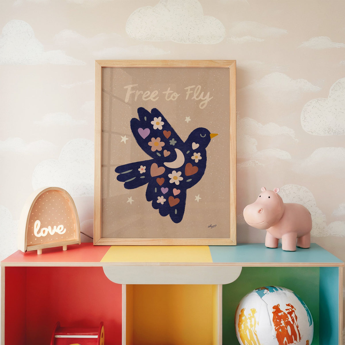 Navy folk bird free to fly nursery print in an oak frame on a rainbow toy shelf against cloud wallpaper