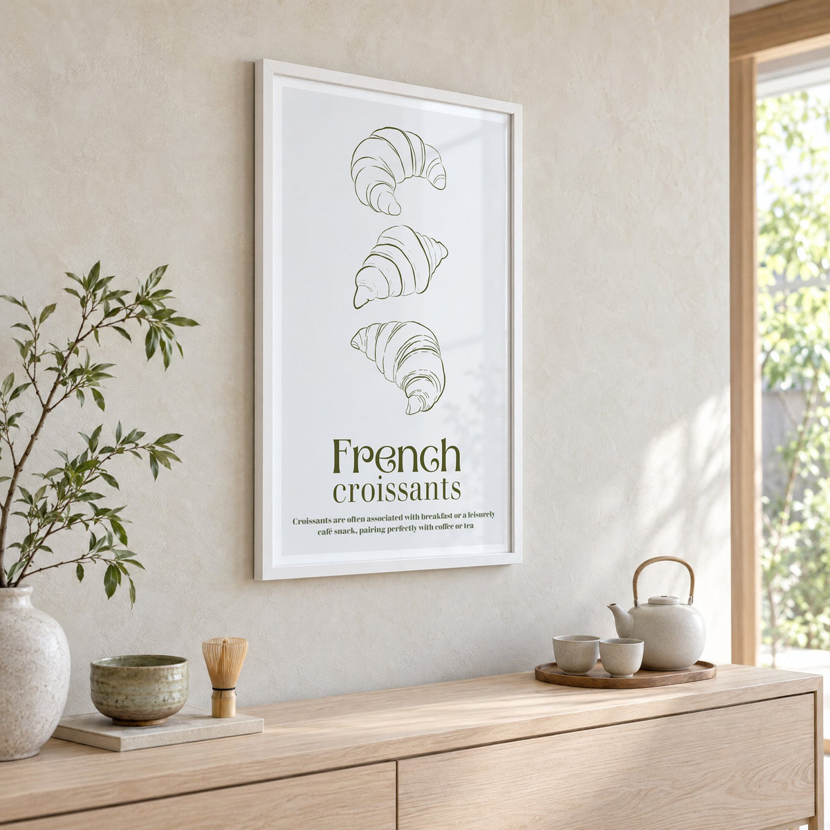 French croissants olive line print in a white frame above a pale wood console with a matcha tea set