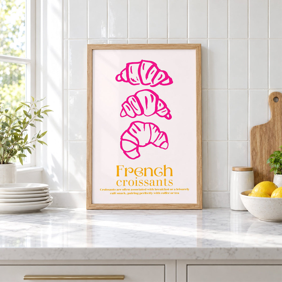French croissants hot pink line print in an oak frame leaning on a marble counter by white tiles and lemons
