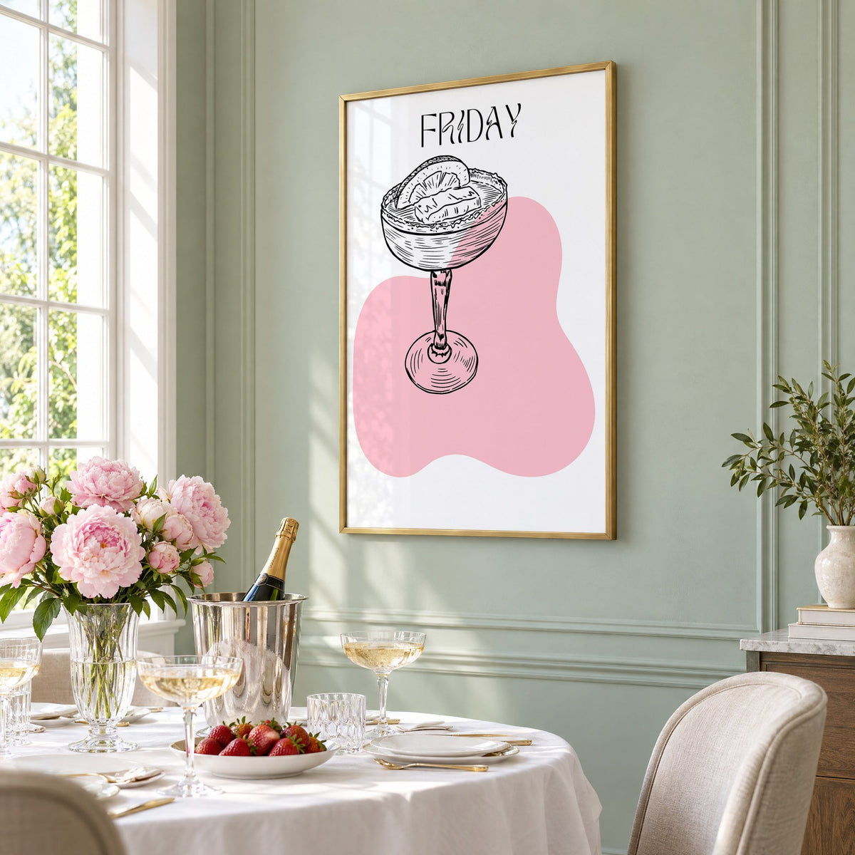FRIDAY engraved coupe cocktail pink blob print in a brass frame on a sage wall above a brunch table with champagne and peonies, seen at an angle