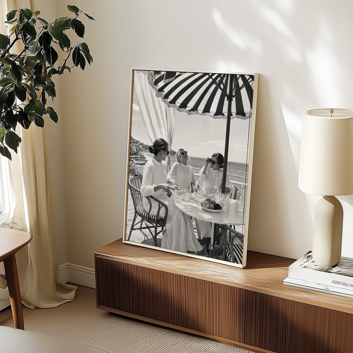 Three friends lunching under a striped umbrella print in a white frame on a walnut sideboard beside a ceramic lamp