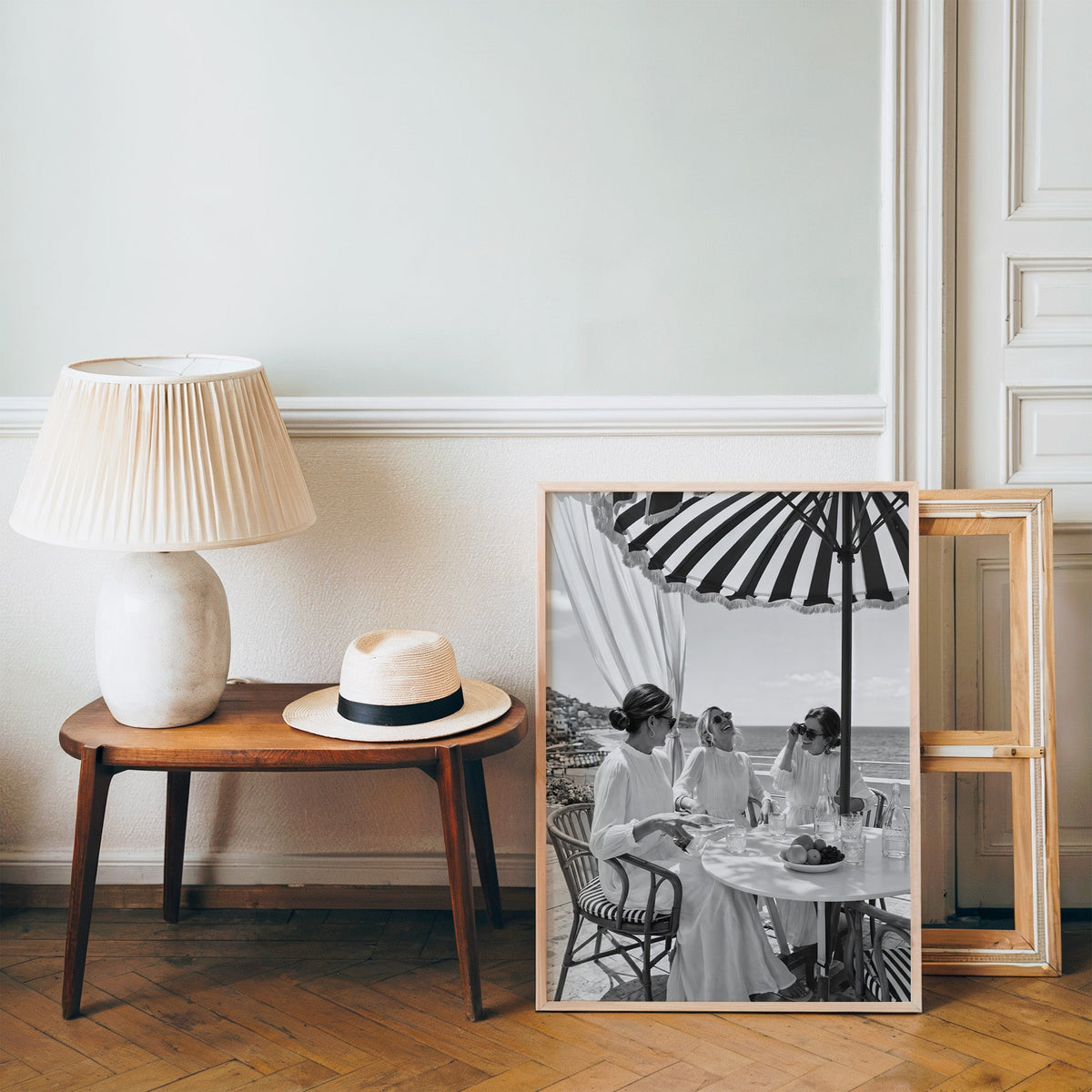 Three friends lunching under a striped umbrella print in a pine frame on a wood floor beside a lamp and straw hat