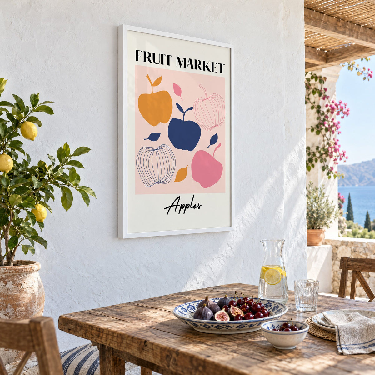 FRUIT MARKET Apples cut-paper print in a white frame on a whitewashed terrace wall above a lunch table with figs and cherries, seen at an angle