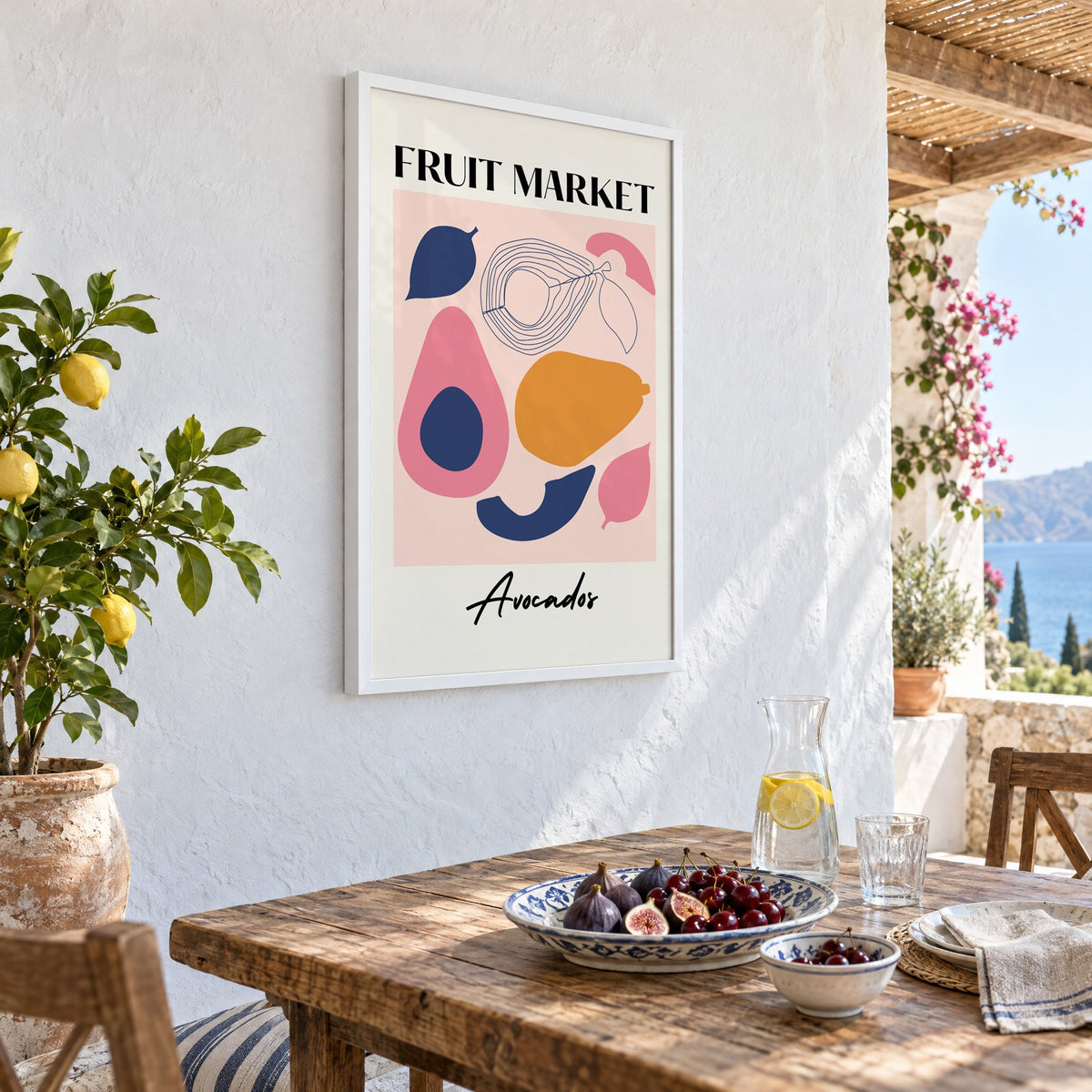 FRUIT MARKET Avocados cut-paper print in a white frame on a whitewashed terrace wall above a lunch table with figs and cherries, seen at an angle