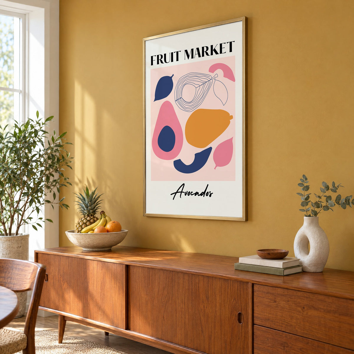 FRUIT MARKET Avocados cut-paper print in a brass frame on a mustard wall above a teak sideboard with a fruit bowl, seen at an angle