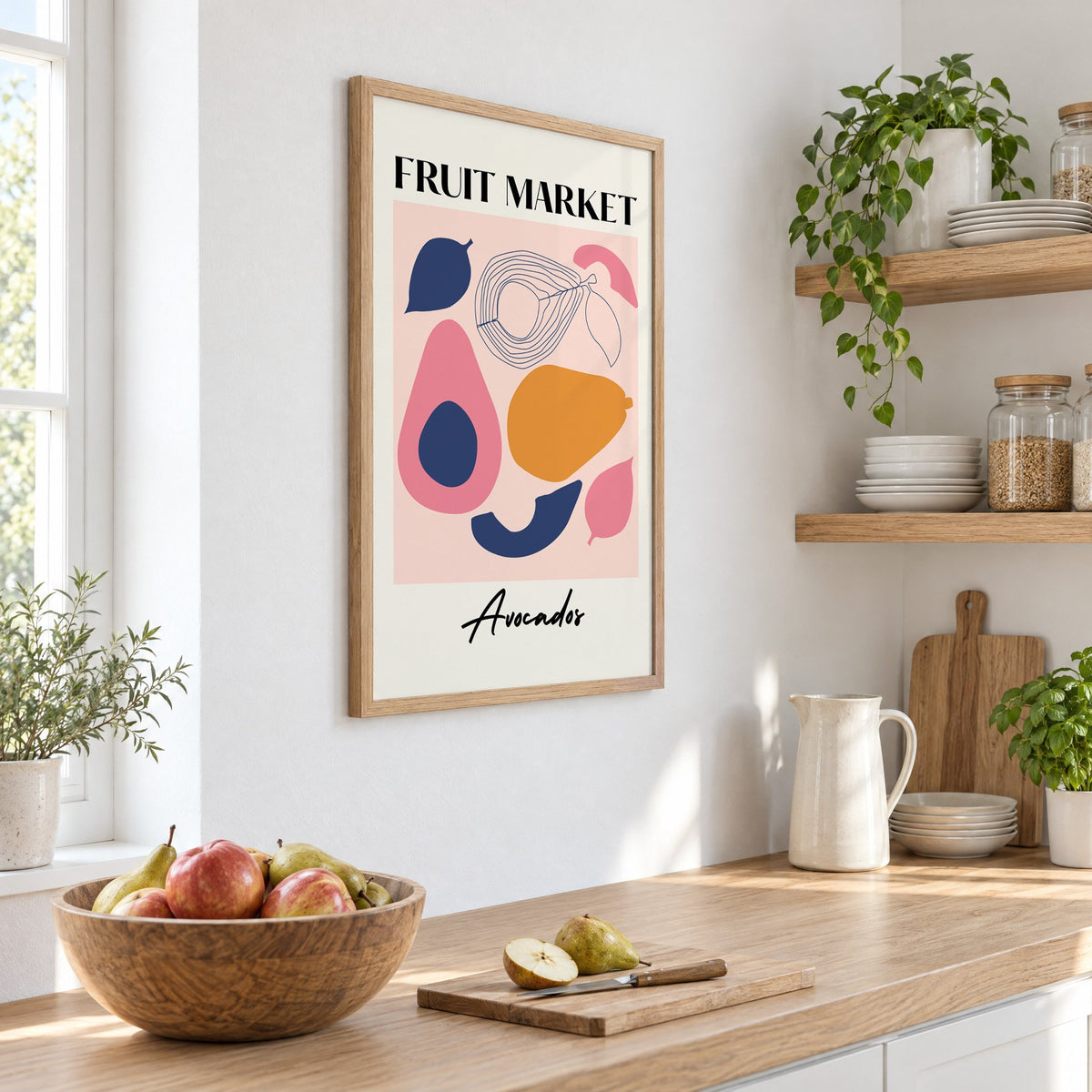 FRUIT MARKET Avocados cut-paper print in an oak frame on a white kitchen wall above a counter with apples and pears, seen at an angle