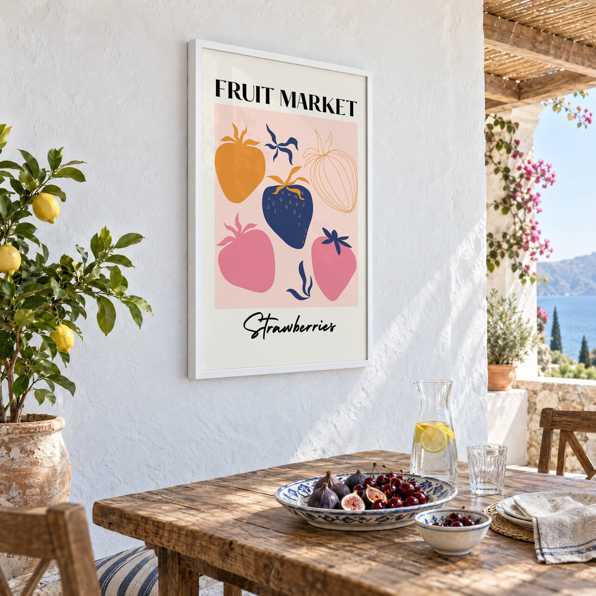 FRUIT MARKET Strawberries cut-paper print in a white frame on a whitewashed terrace wall above a lunch table with figs and cherries, seen at an angle