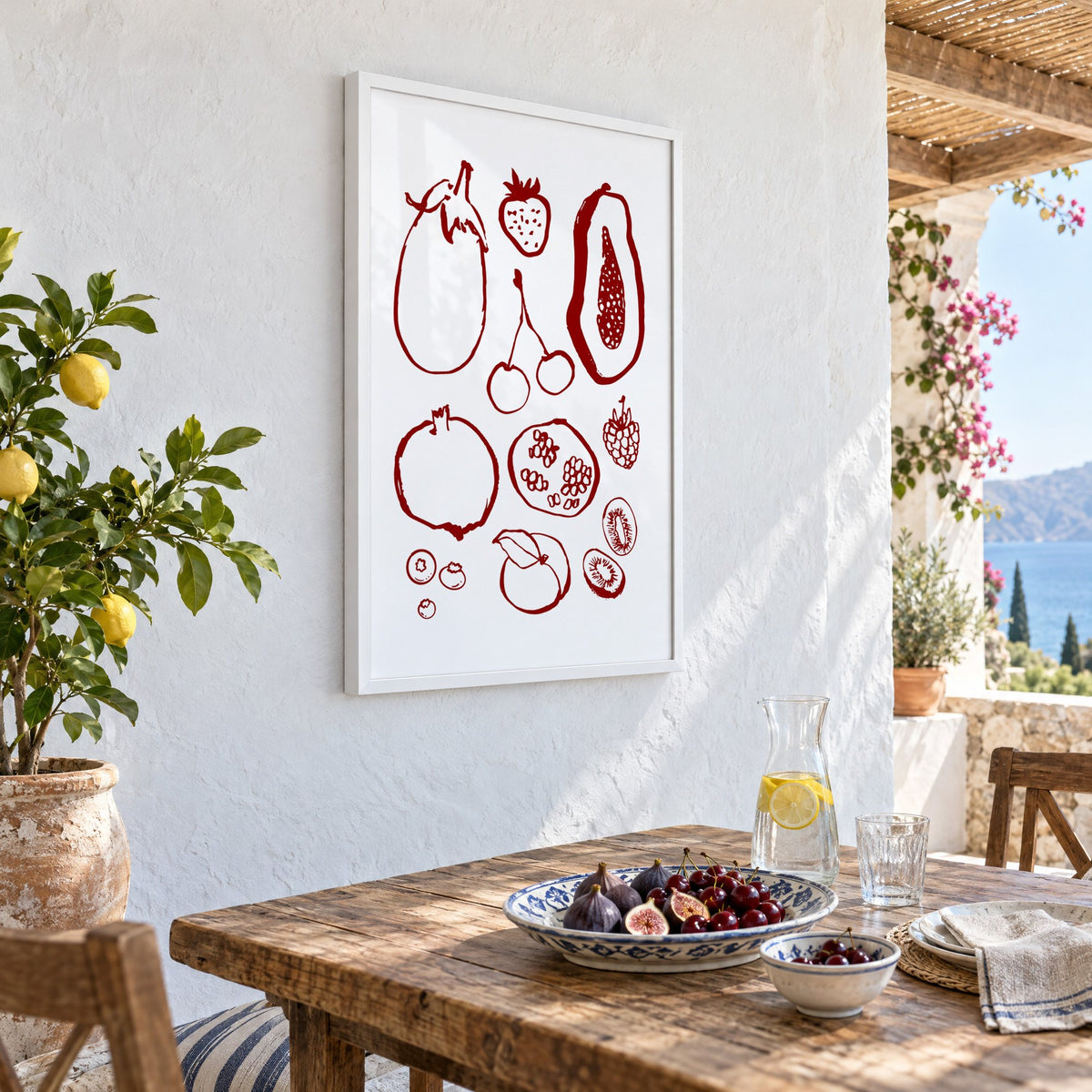 Burgundy ink fruit sketch pattern print in a white frame on a whitewashed terrace wall above a lunch table with figs and cherries, seen at an angle