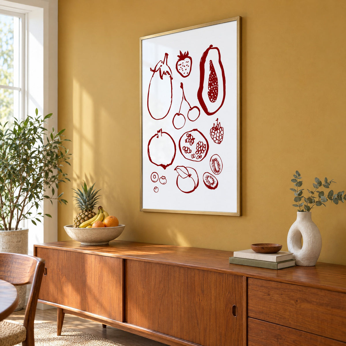 Burgundy ink fruit sketch pattern print in a brass frame on a mustard wall above a teak sideboard with a fruit bowl, seen at an angle