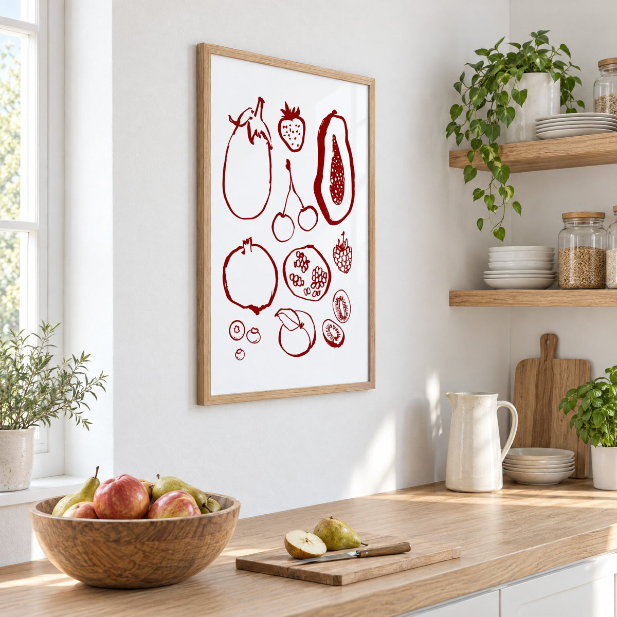 Burgundy ink fruit sketch pattern print in an oak frame on a white kitchen wall above a counter with apples and pears, seen at an angle