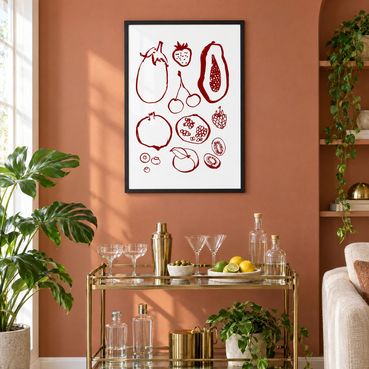 Burgundy ink fruit sketch pattern print in a black frame on a terracotta wall above a brass bar cart with coupes and citrus