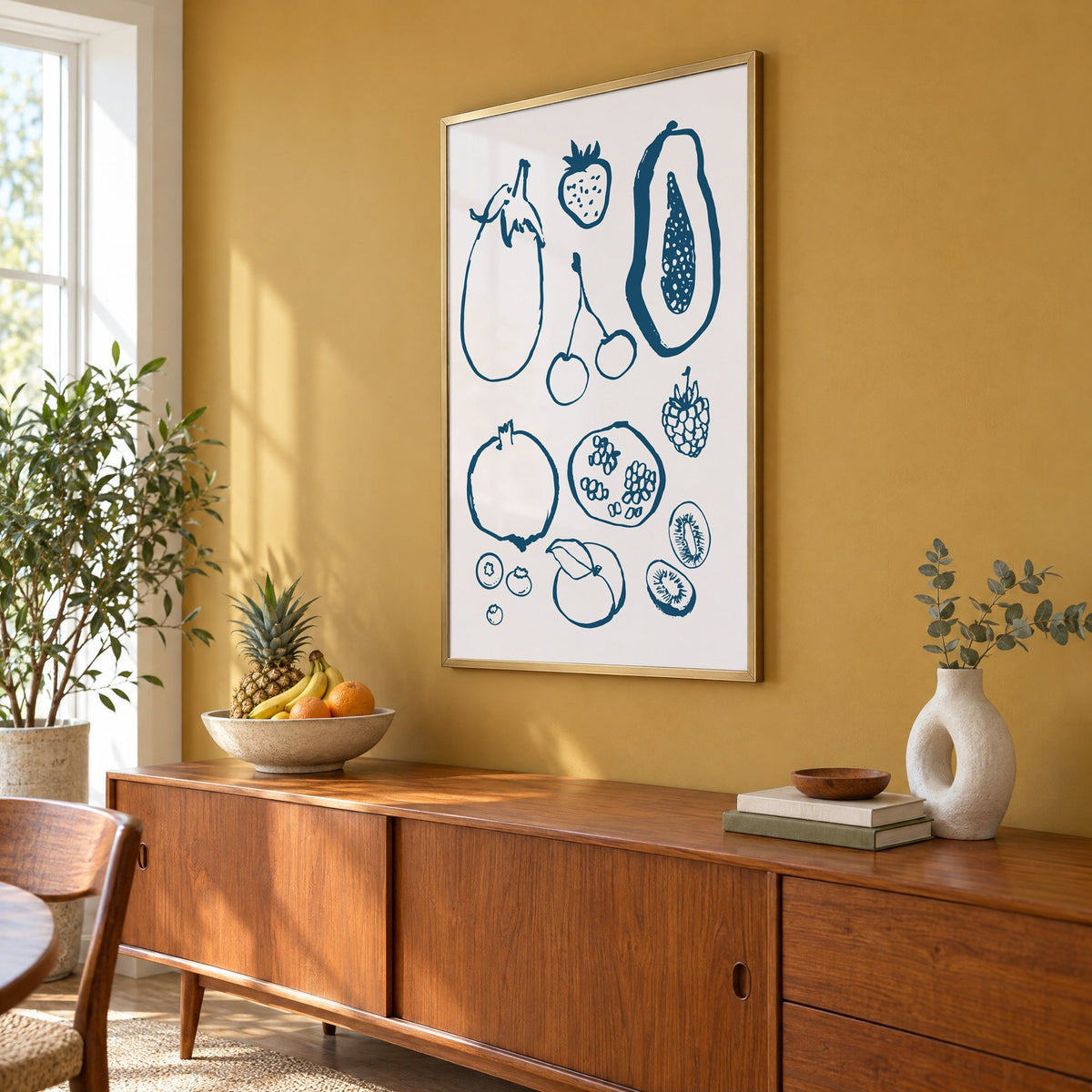petrol blue ink fruit sketch pattern print in a brass frame on a mustard wall above a teak sideboard with a fruit bowl, seen at an angle
