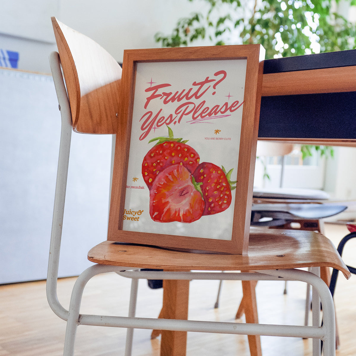Fruit Yes Please strawberries print in a walnut frame resting on a plywood studio chair beside a dark desk