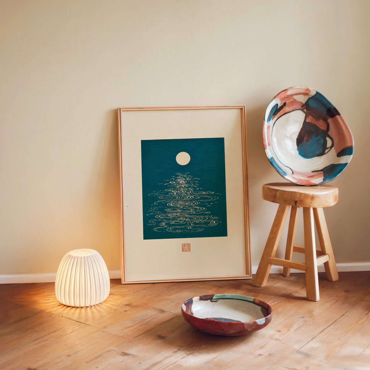 full moon over teal water print in a light wood frame leaning on a wooden floor by a glowing lamp and stool