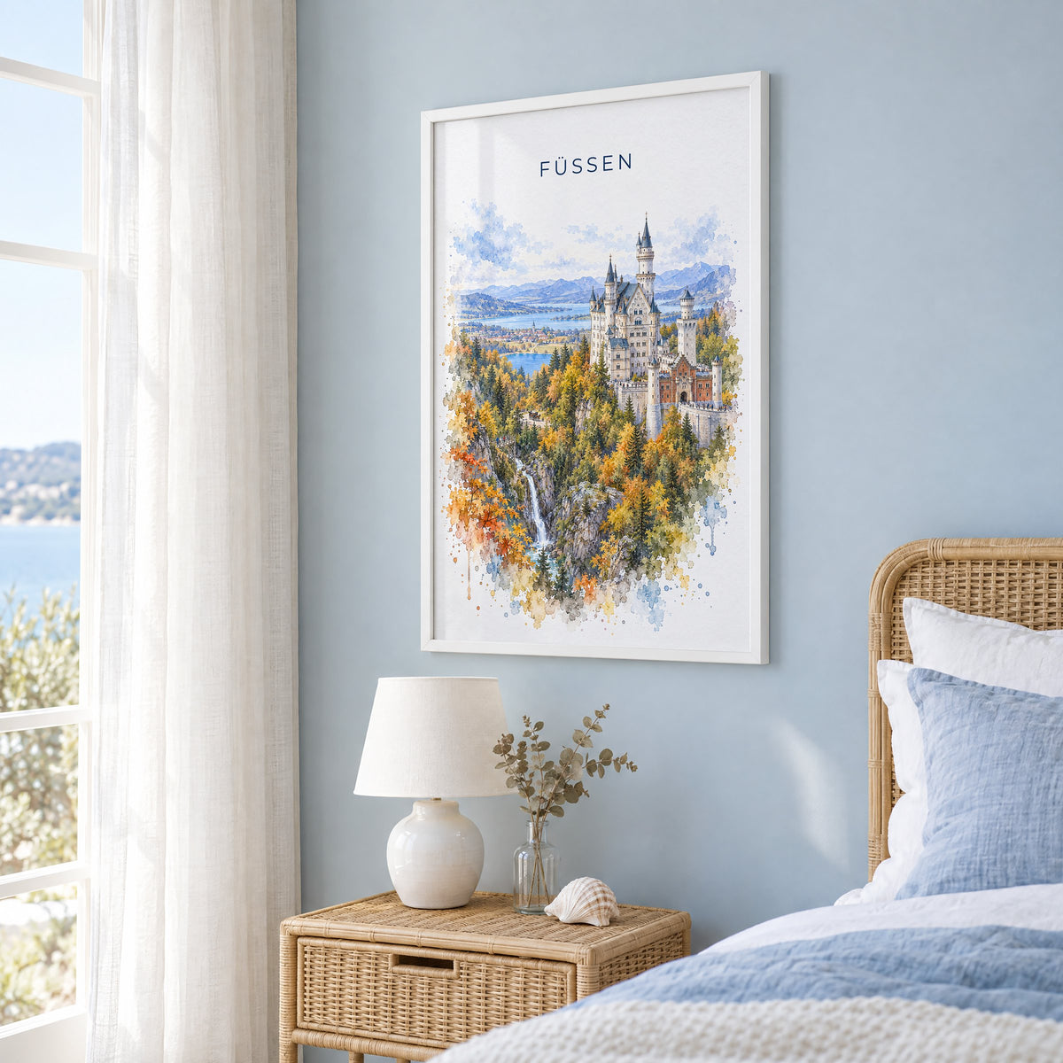 Füssen watercolor print in a white frame on a pale blue wall beside a rattan bed in a coastal bedroom