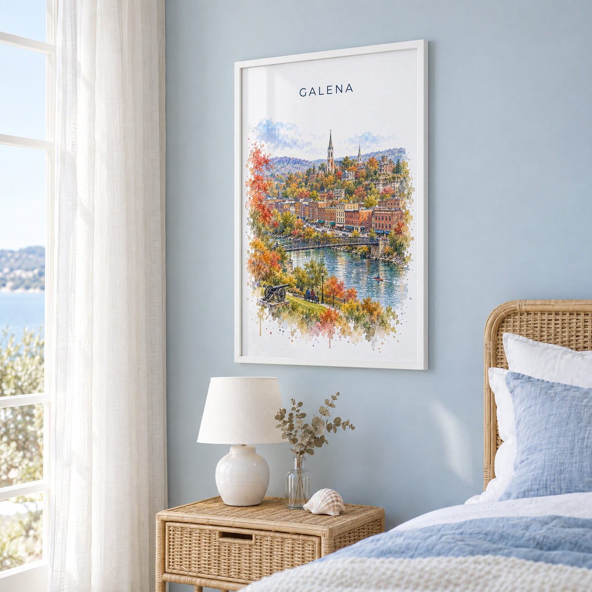 Galena, Illinois watercolor print in a white frame on a pale blue wall beside a rattan bed in a coastal bedroom