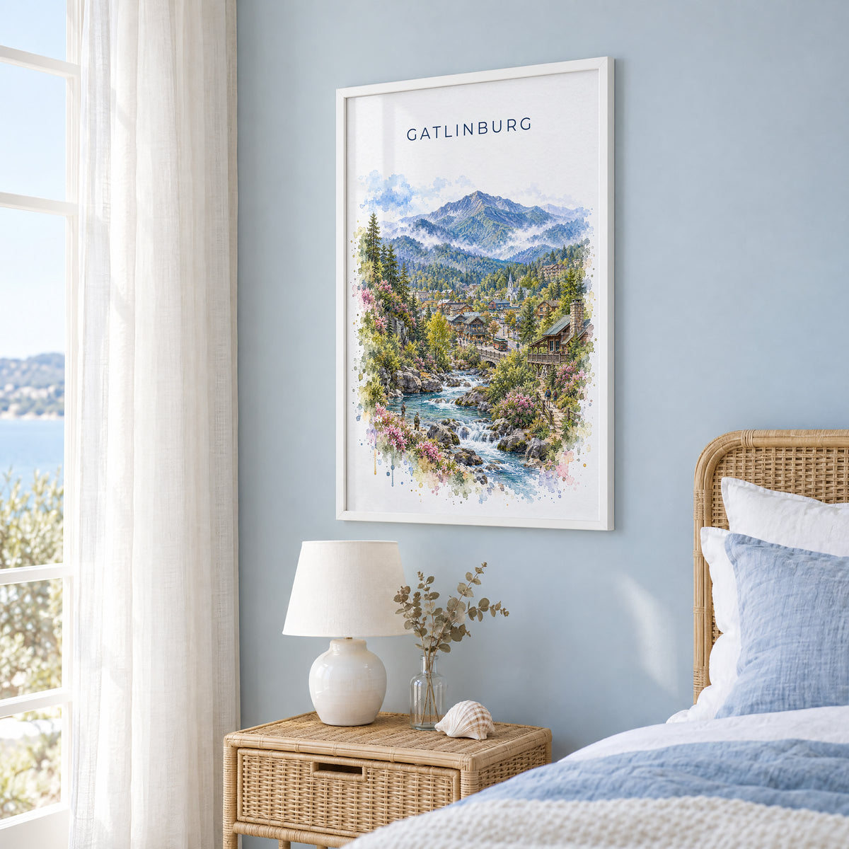 Gatlinburg watercolor print in a white frame on a pale blue wall beside a rattan bed in a coastal bedroom