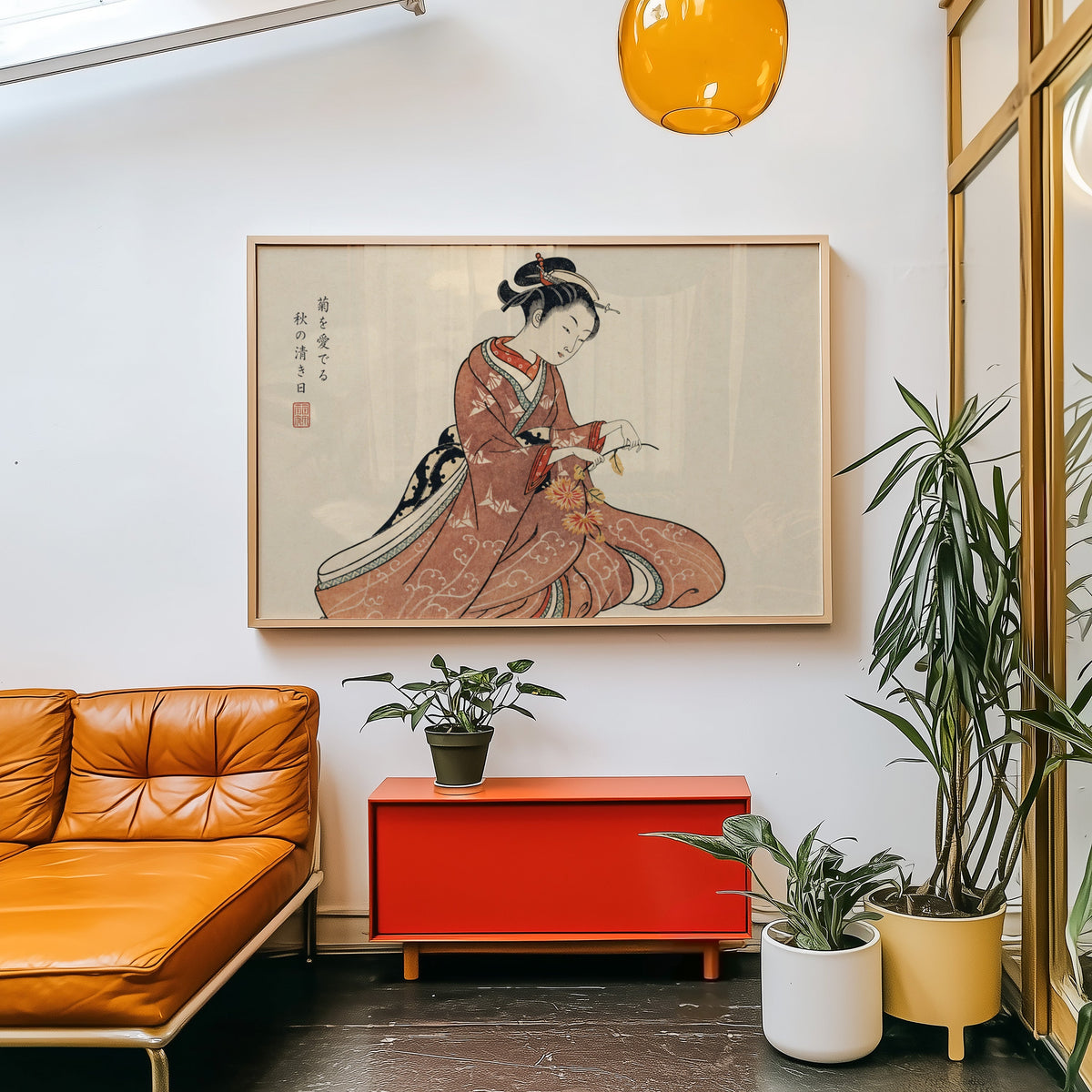 Kimono woman holding chrysanthemums print in a light wood frame above a red cabinet beside a tan leather daybed
