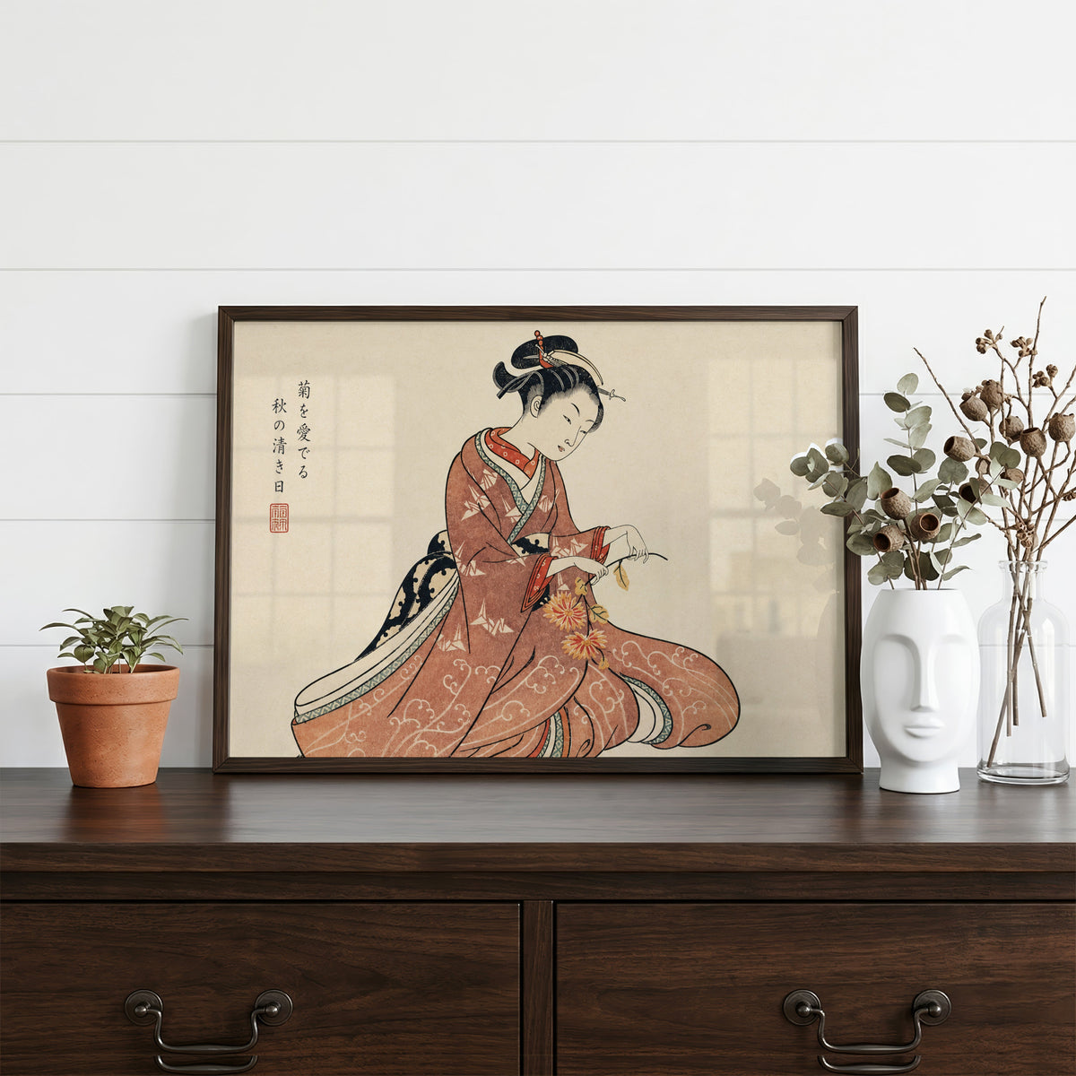 Kimono woman holding chrysanthemums print in a dark walnut frame on a dark wood dresser beside a white face vase