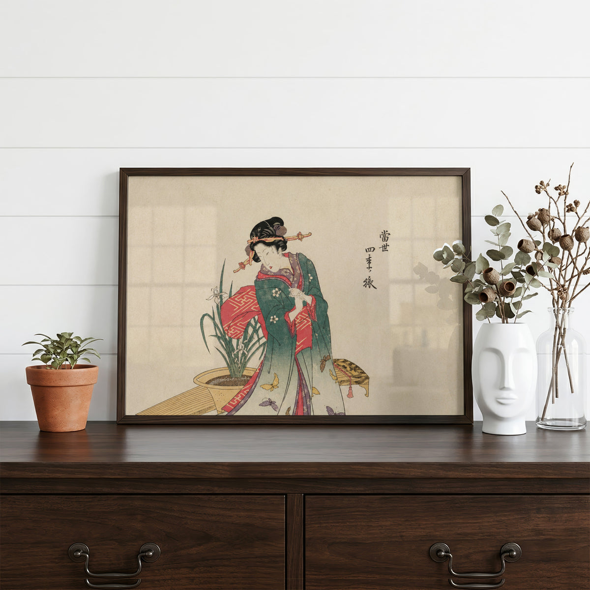 Woman in green kimono with butterflies print in a dark walnut frame on a dark wood dresser beside a white face vase