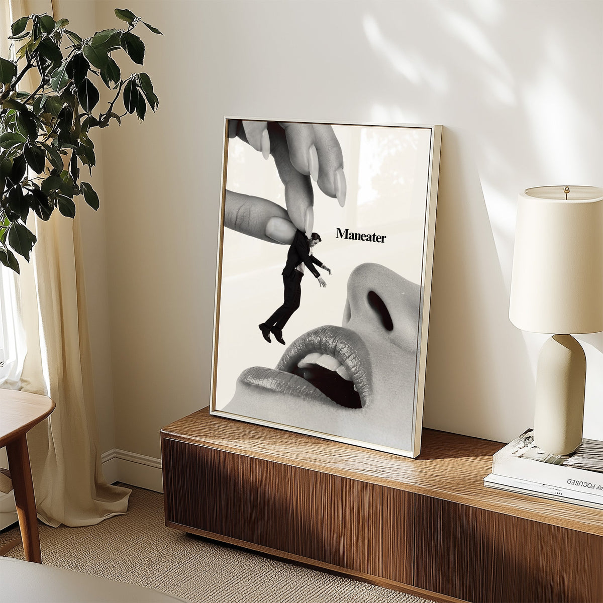 Giant hand lifting tiny suited man collage print in a white frame on a walnut sideboard beside a ceramic lamp