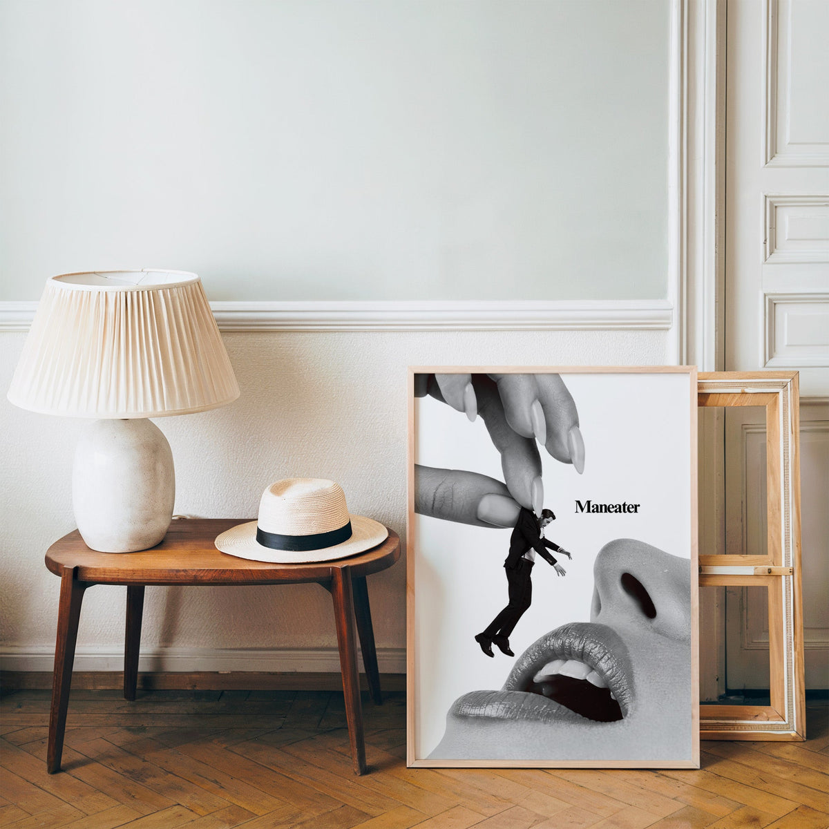Giant hand lifting tiny suited man collage print in a pine frame on a wood floor beside a lamp and straw hat