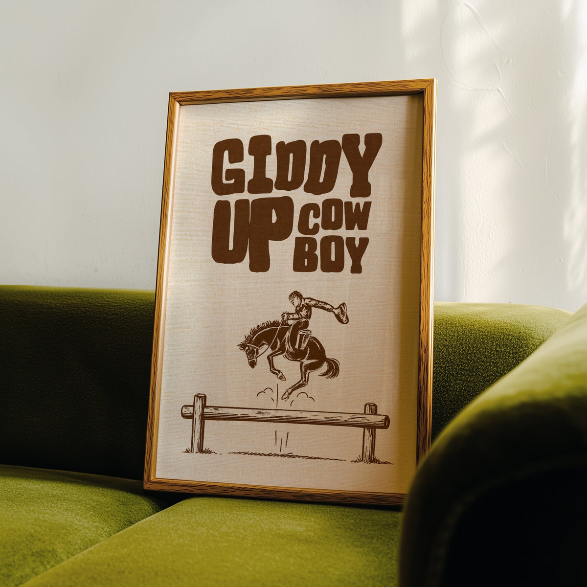 Giddy Up Cowboy cream print in an oak frame leaning on a green velvet sofa
