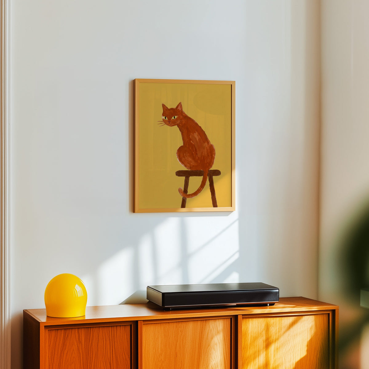 Mustard ginger cat print in a light oak frame above a teak sideboard with a yellow lamp