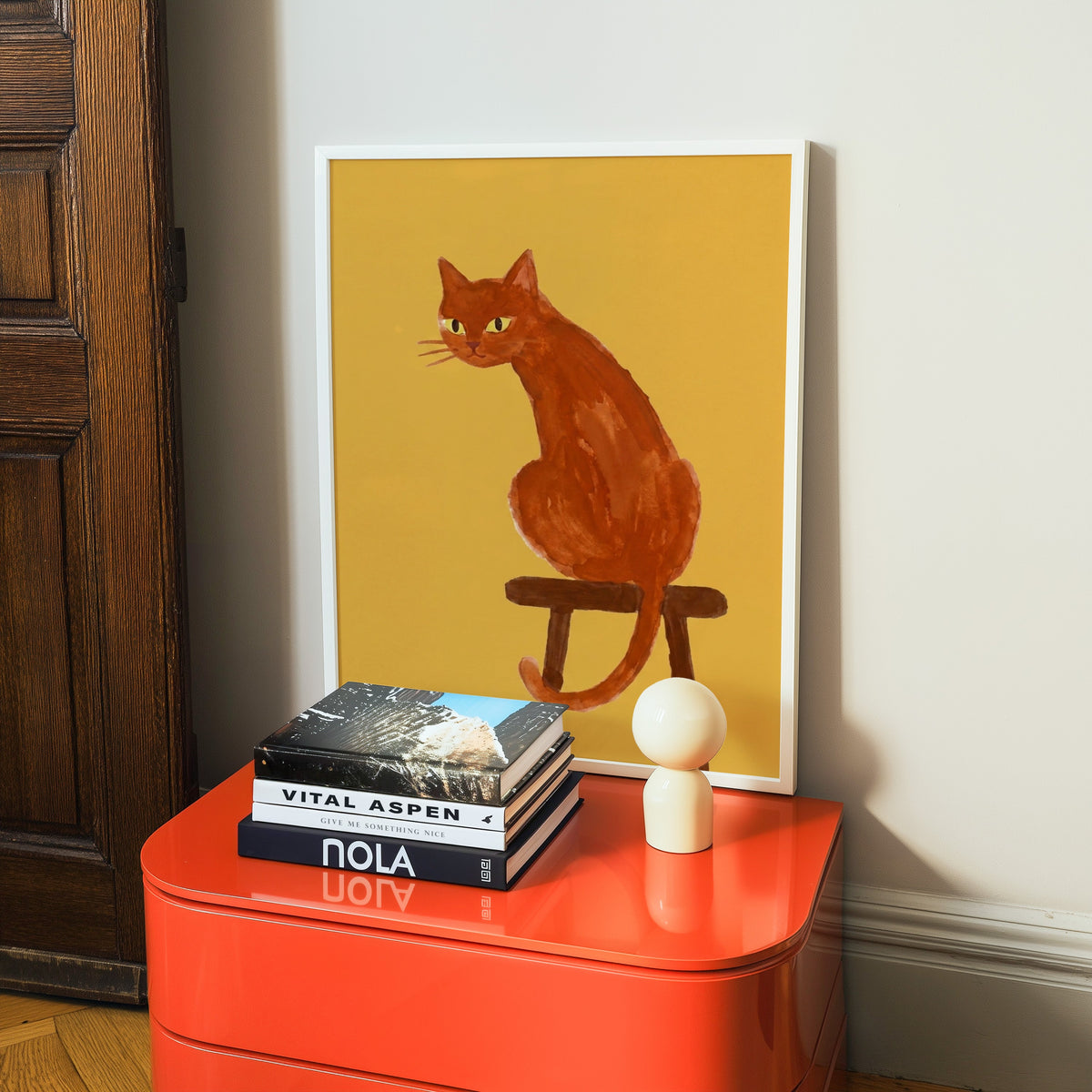Orange cat watercolor print in a white frame on a glossy orange cabinet with books