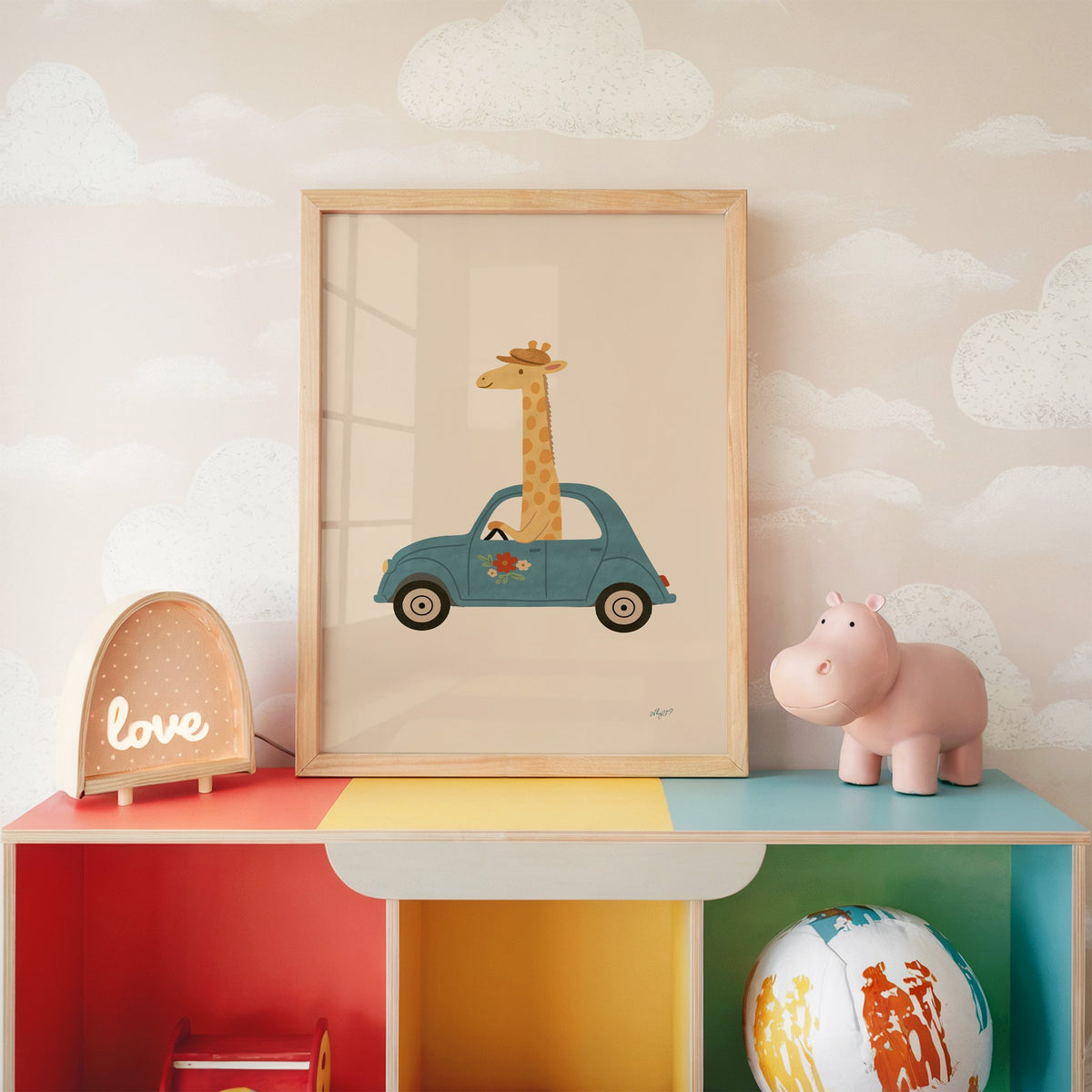 Giraffe driving a teal car nursery print in an oak frame on a rainbow toy shelf against cloud wallpaper