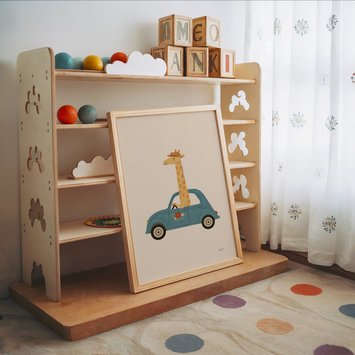 Giraffe driving a teal car nursery print in an oak frame leaning on a toy shelf with wooden letter blocks