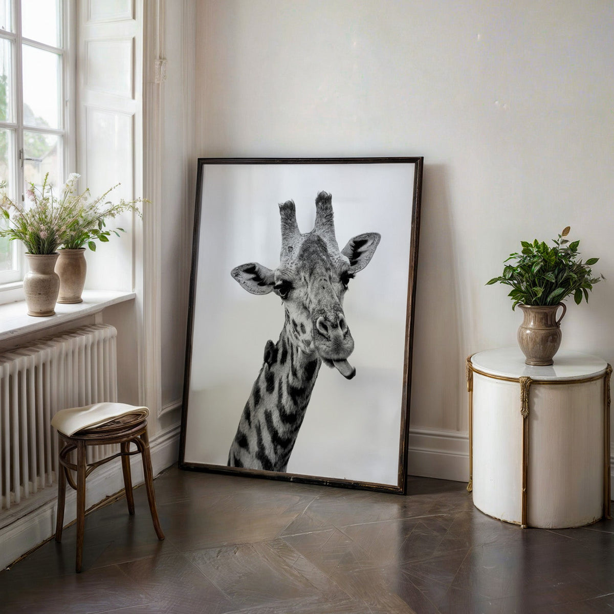 Giraffe sticking out tongue with small bird print in a black frame on a dark stone floor beside a white side table