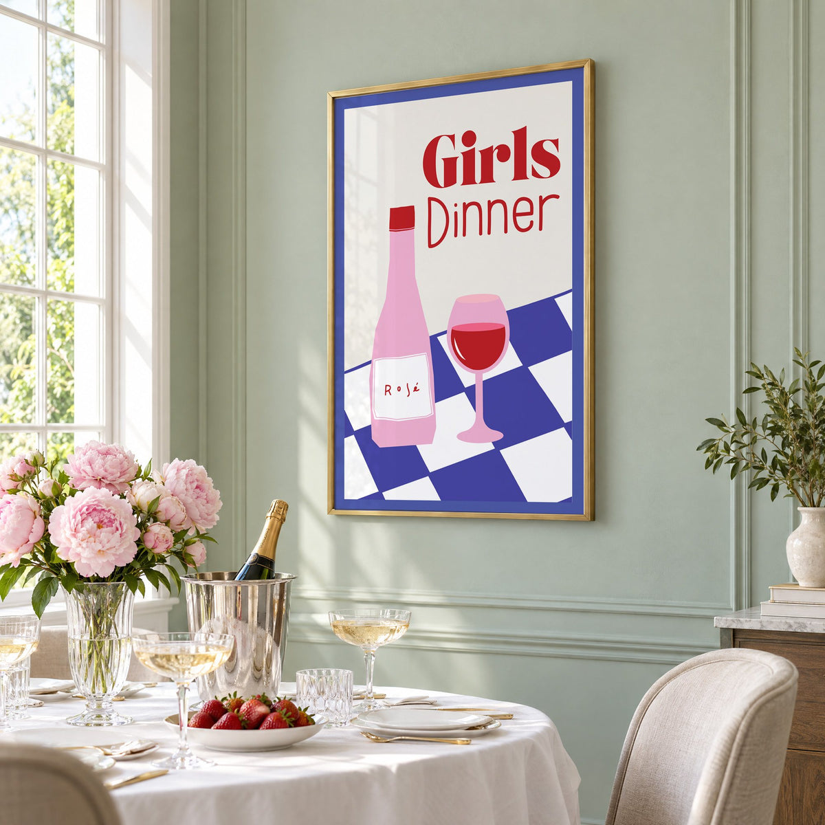 Girls Dinner rosé bottle on checkered table print in a brass frame on a sage wall above a brunch table with champagne and peonies, seen at an angle