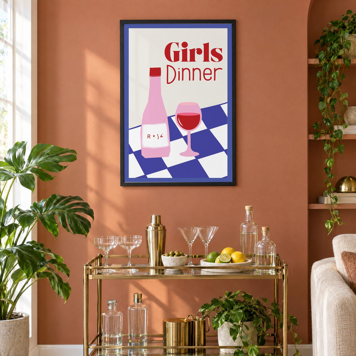 Girls Dinner rosé bottle on checkered table print in a black frame on a terracotta wall above a brass bar cart with coupes and citrus