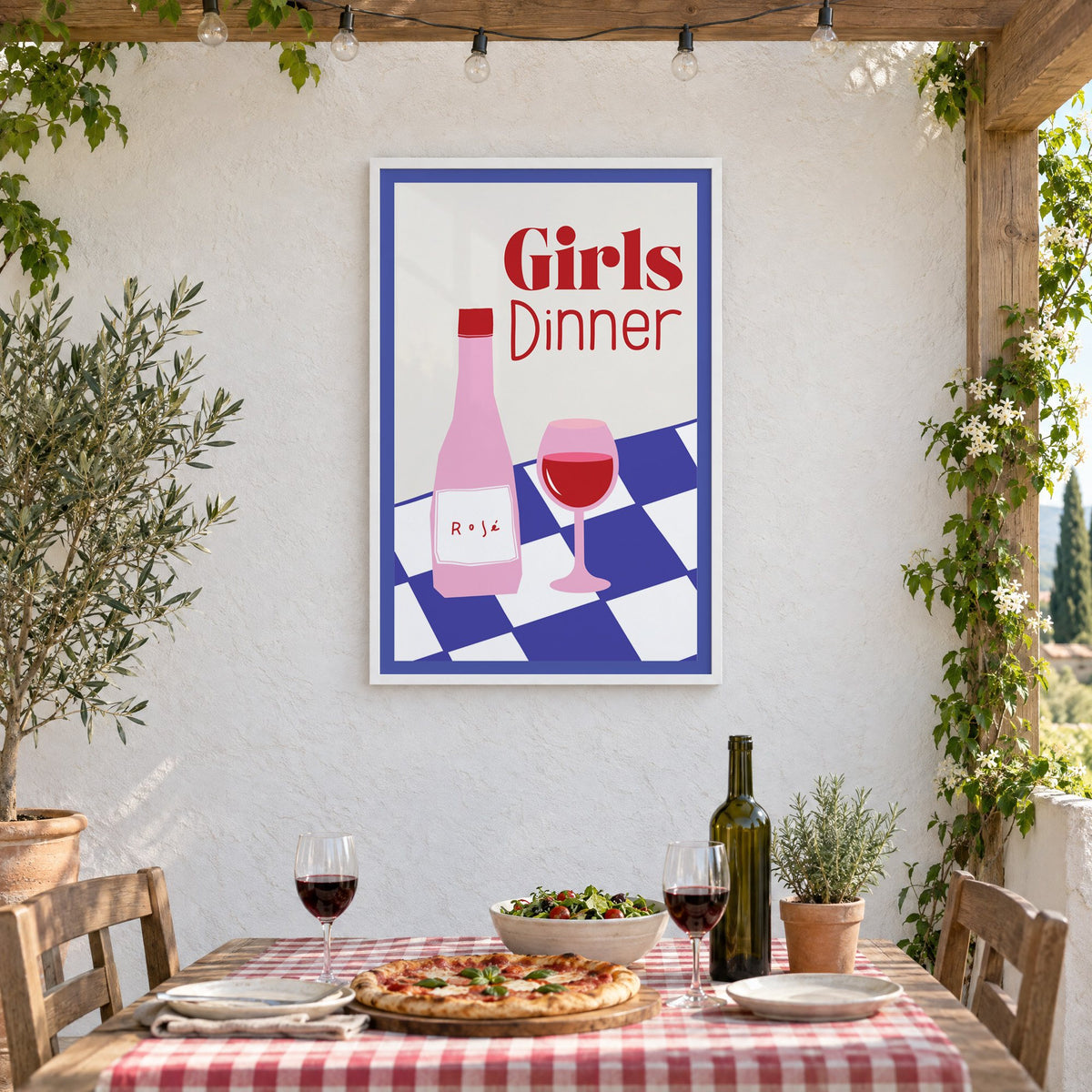 Girls Dinner rosé bottle on checkered table print in a white frame on a stucco wall above a checked-cloth table with pizza and red wine