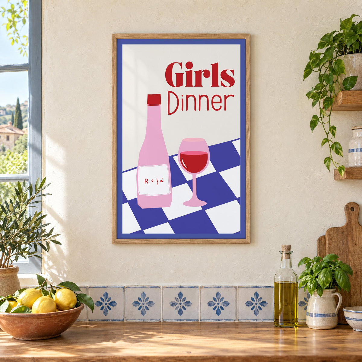 Girls Dinner rosé bottle on checkered table print in an oak frame on a cream plaster wall above a worktop with a bowl of lemons and blue-and-white tiles
