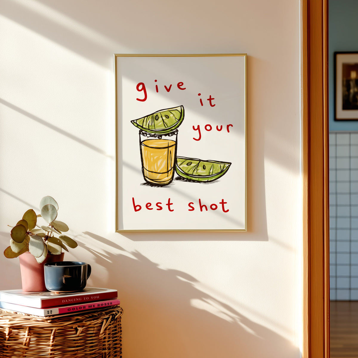 Tequila shot print shown in a gold frame above a basket of books and a pilea plant