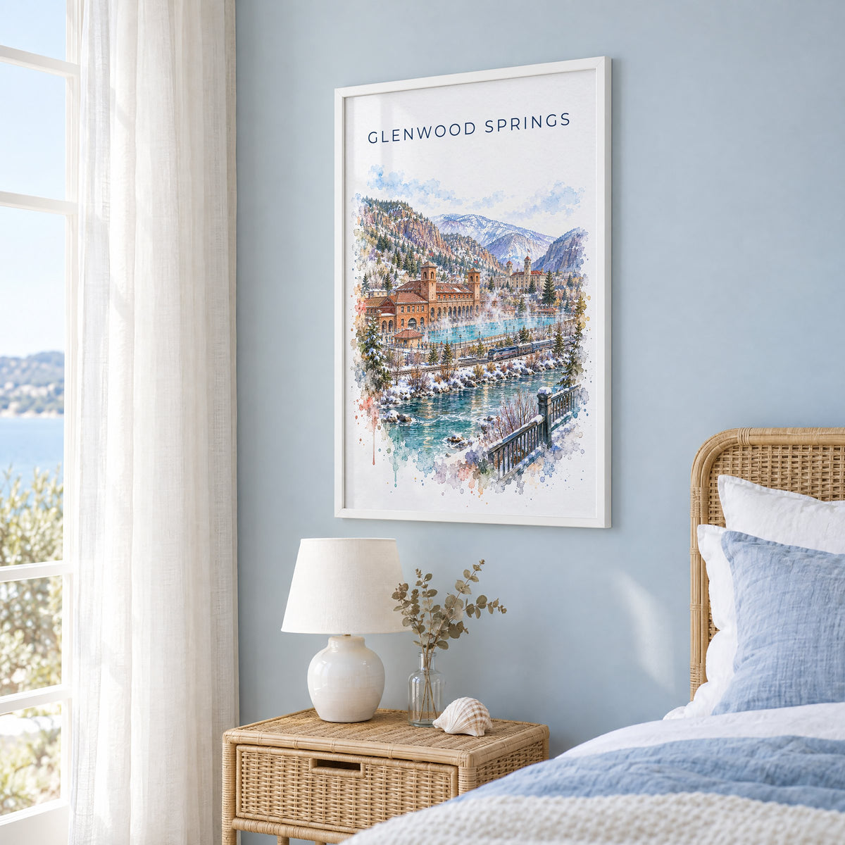 Glenwood Springs watercolor print in a white frame on a pale blue wall beside a rattan bed in a coastal bedroom