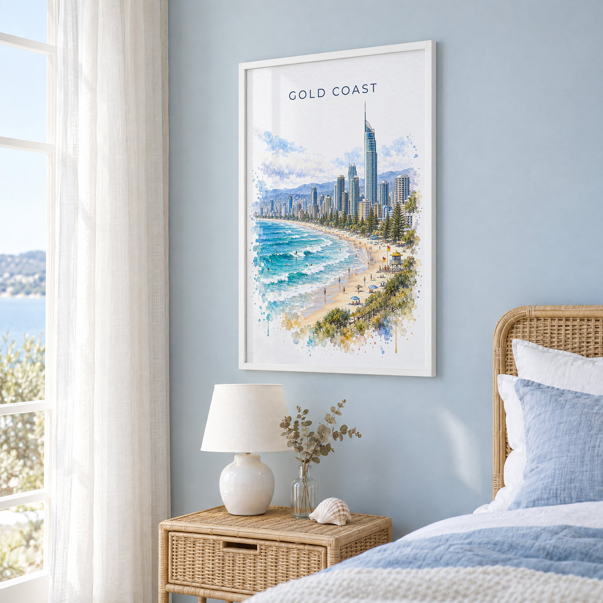 Gold Coast watercolor print in a white frame on a pale blue wall beside a rattan bed in a coastal bedroom